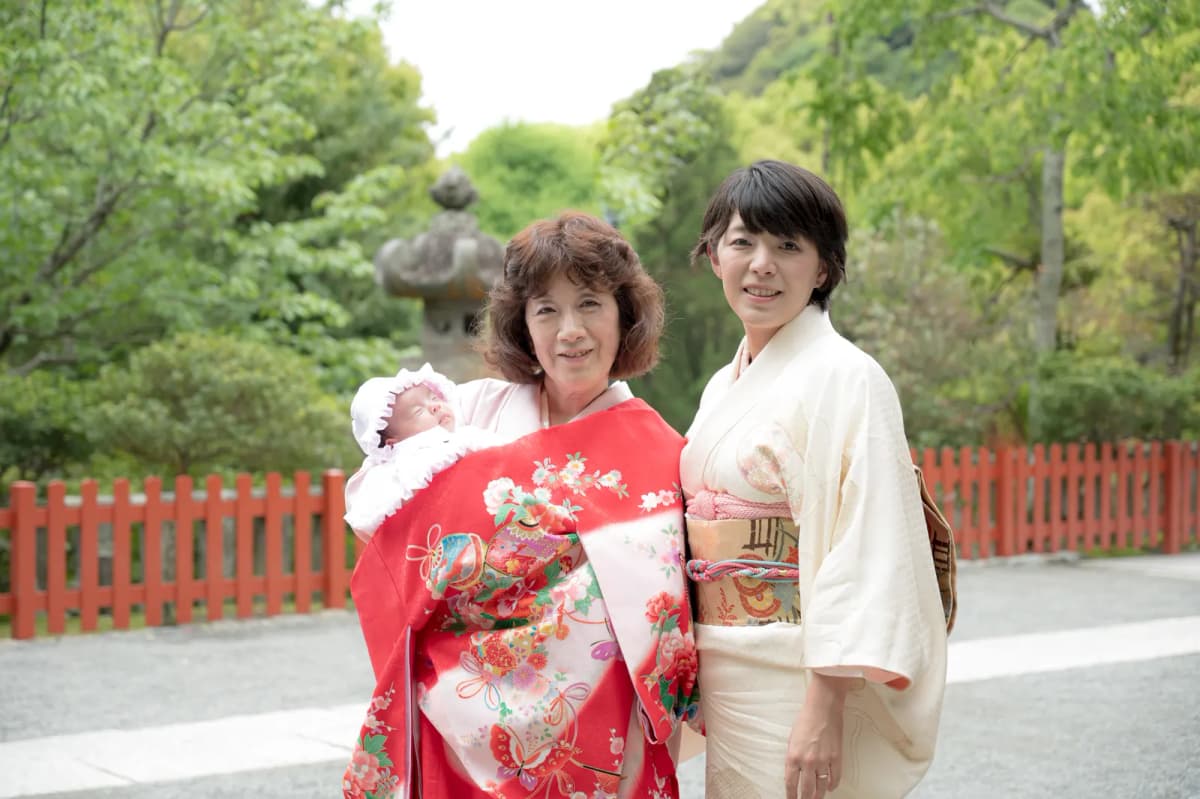 神社の境内で祝い着の赤ちゃんを抱く祖母と母親
