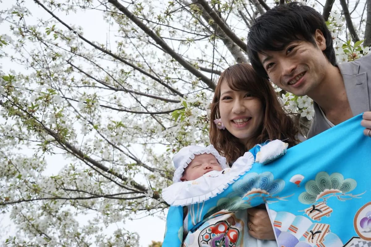 桜の下で祝い着の赤ちゃんを抱く両親
